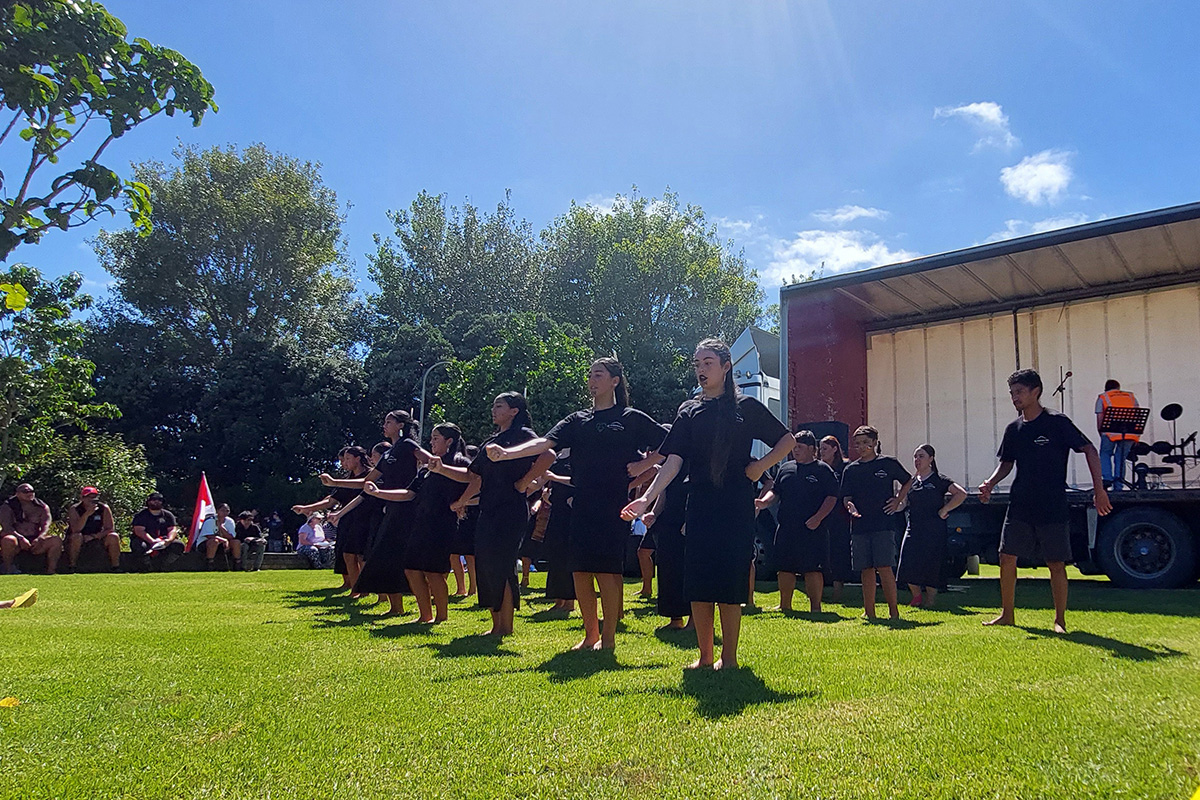 A group of children dressed in black perform kapa haka on grass in front of a stage.